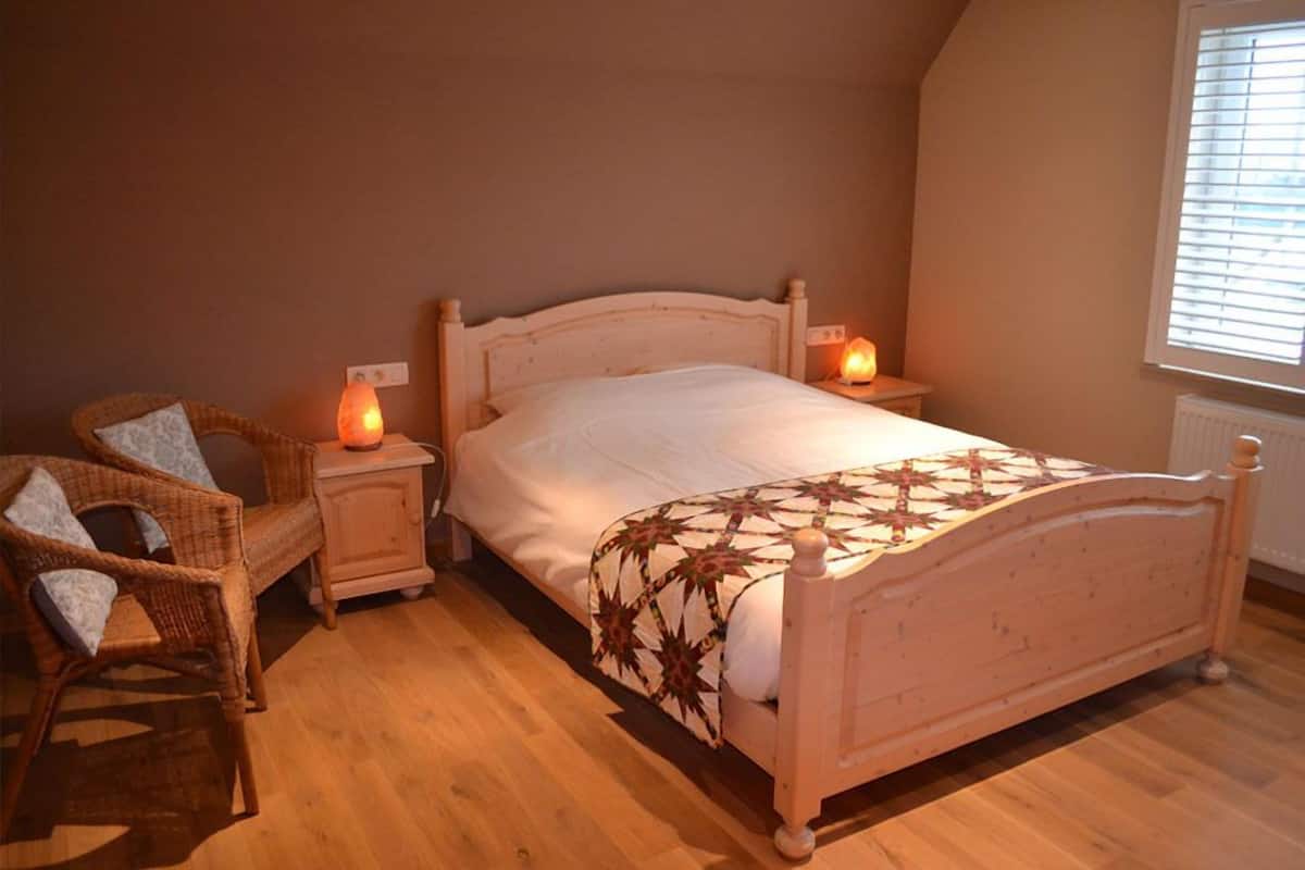 A bedroom with a wooden bed, two bedside tables, a wicker chair, and a window with blinds.