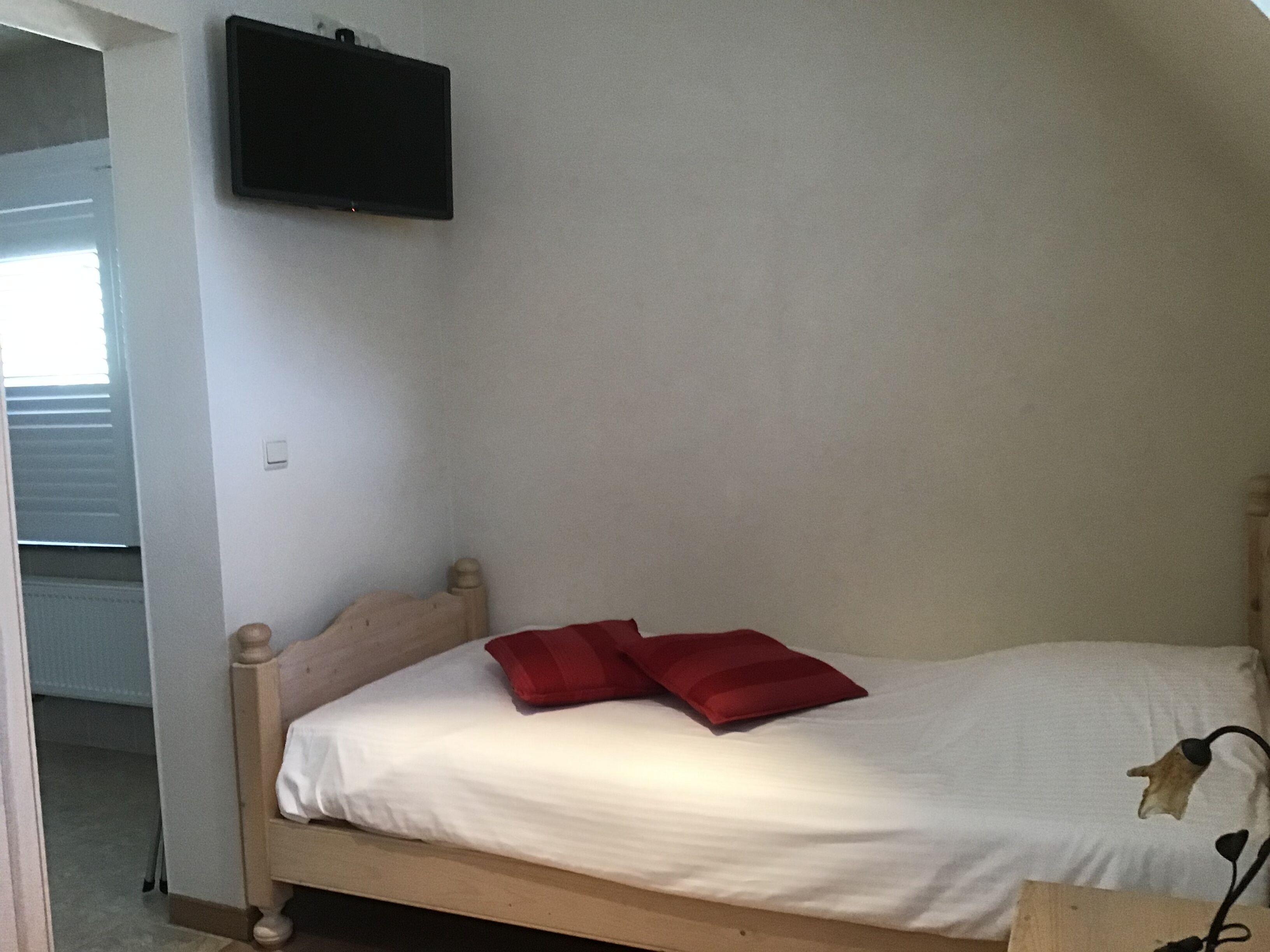 Una cama individual con estructura de madera, ropa de cama blanca y dos almohadas rojas. Un televisor de pantalla plana está montado en la pared sobre la cama.