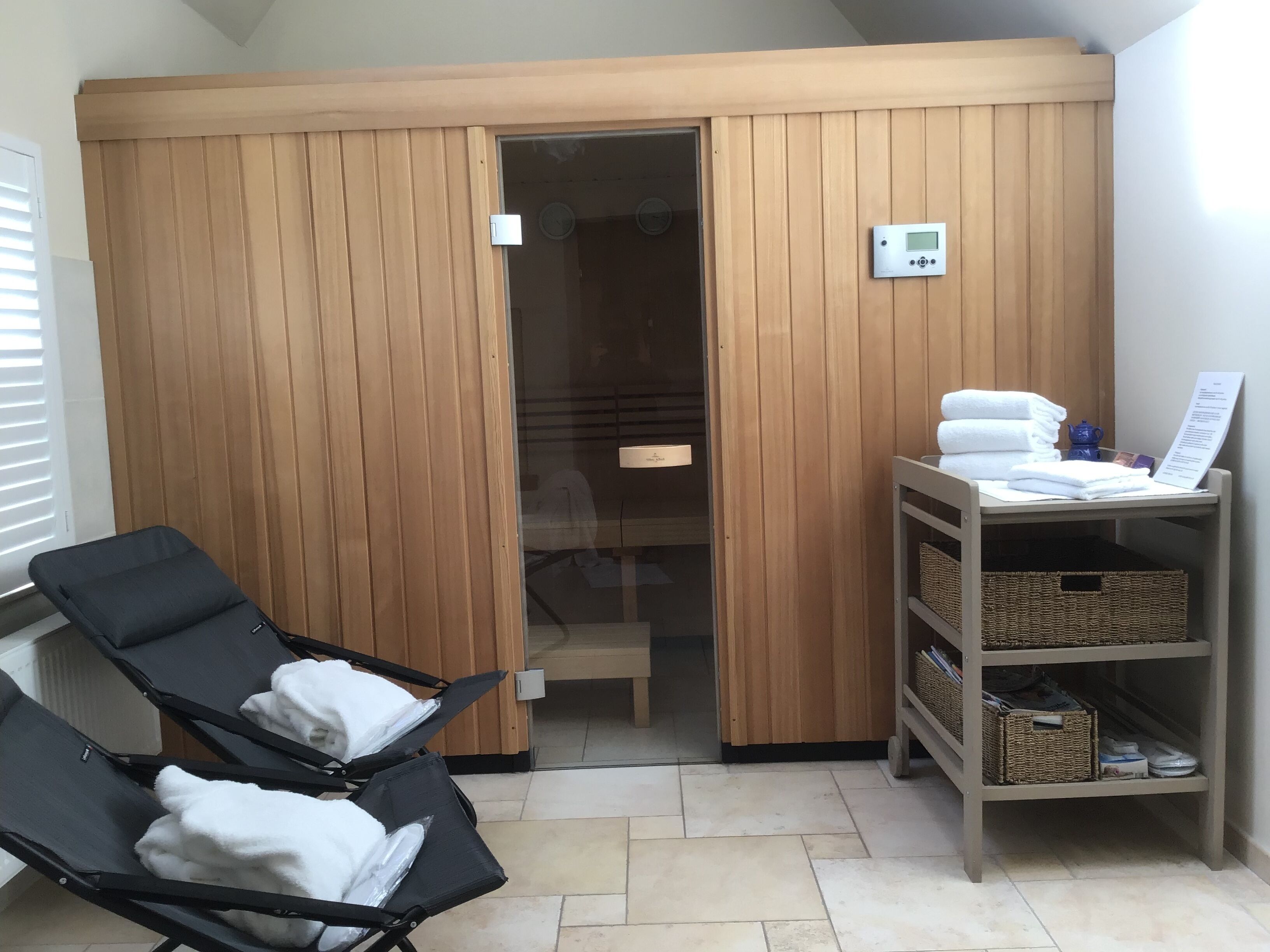 A room with a wooden sliding door, two reclining chairs with towels, a small table with towels and a menu, and a wicker basket shelf.