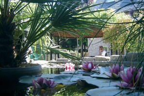 A tranquil garden with water lilies, a gazebo, and a brick building in the background. - Wisteria (Herne)