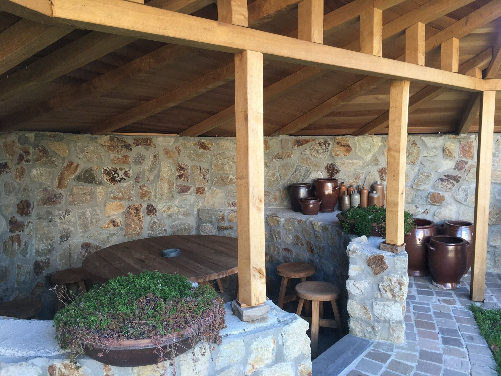 Un muro de piedra rústico con una mesa y taburetes de madera, rodeado de pilares de piedra y grandes macetas de cerámica.