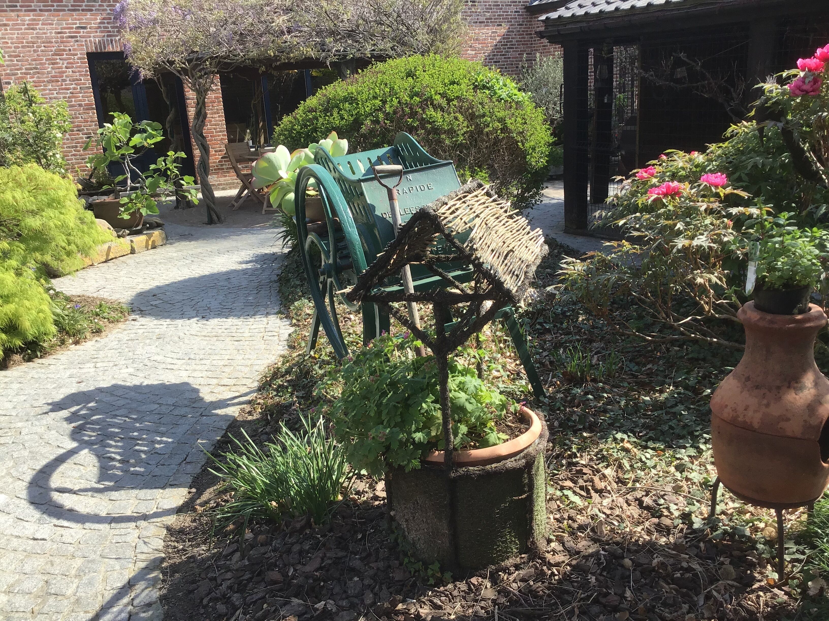 A garden pathway with a rustic water pump, potted plants, and a brick building in the background.