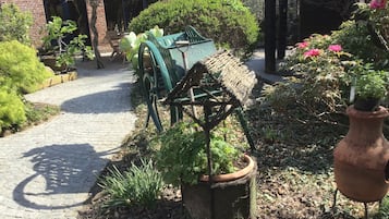 A garden pathway with a rustic water pump, potted plants, and a brick building in the background.