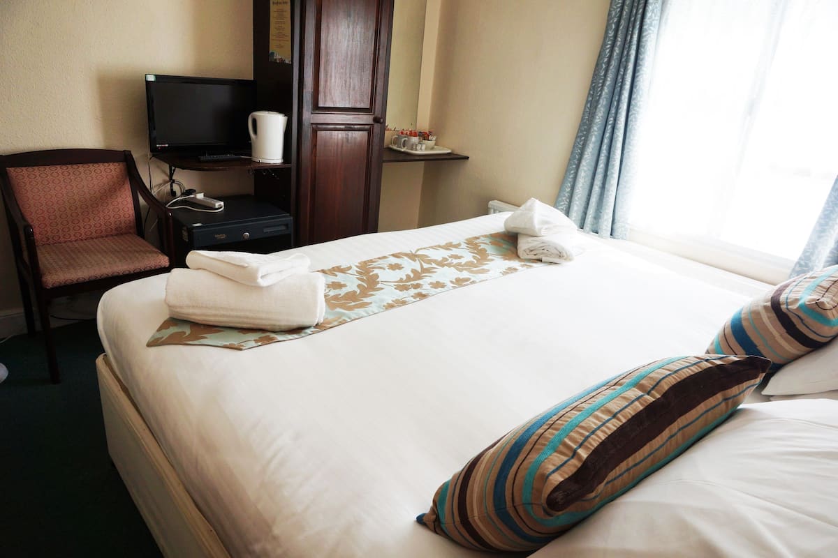 Una habitación de hotel con cama, una silla, televisor, cafetera y ventana con cortinas.