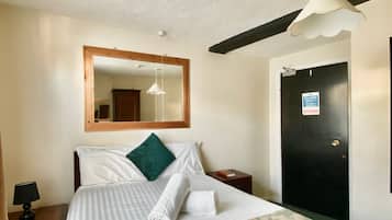 A neatly made bed with white linens and a green pillow, a bedside table with a lamp, a wooden-framed mirror above the bed, and a black door with a sign.