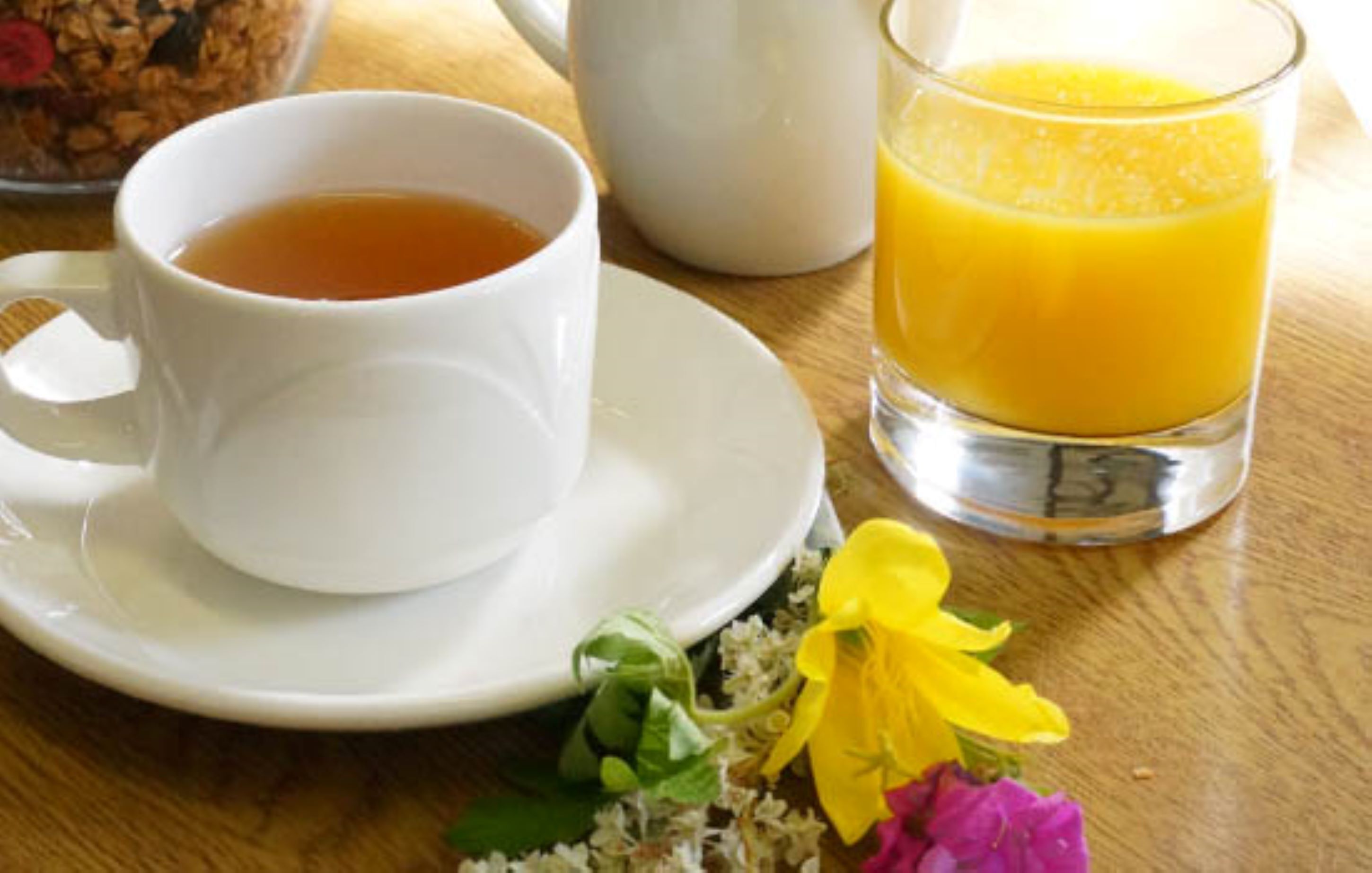 Uma chávena branca de chá sobre um pires, um copo de sumo de laranja e um pequeno buquê de flores numa mesa de madeira.