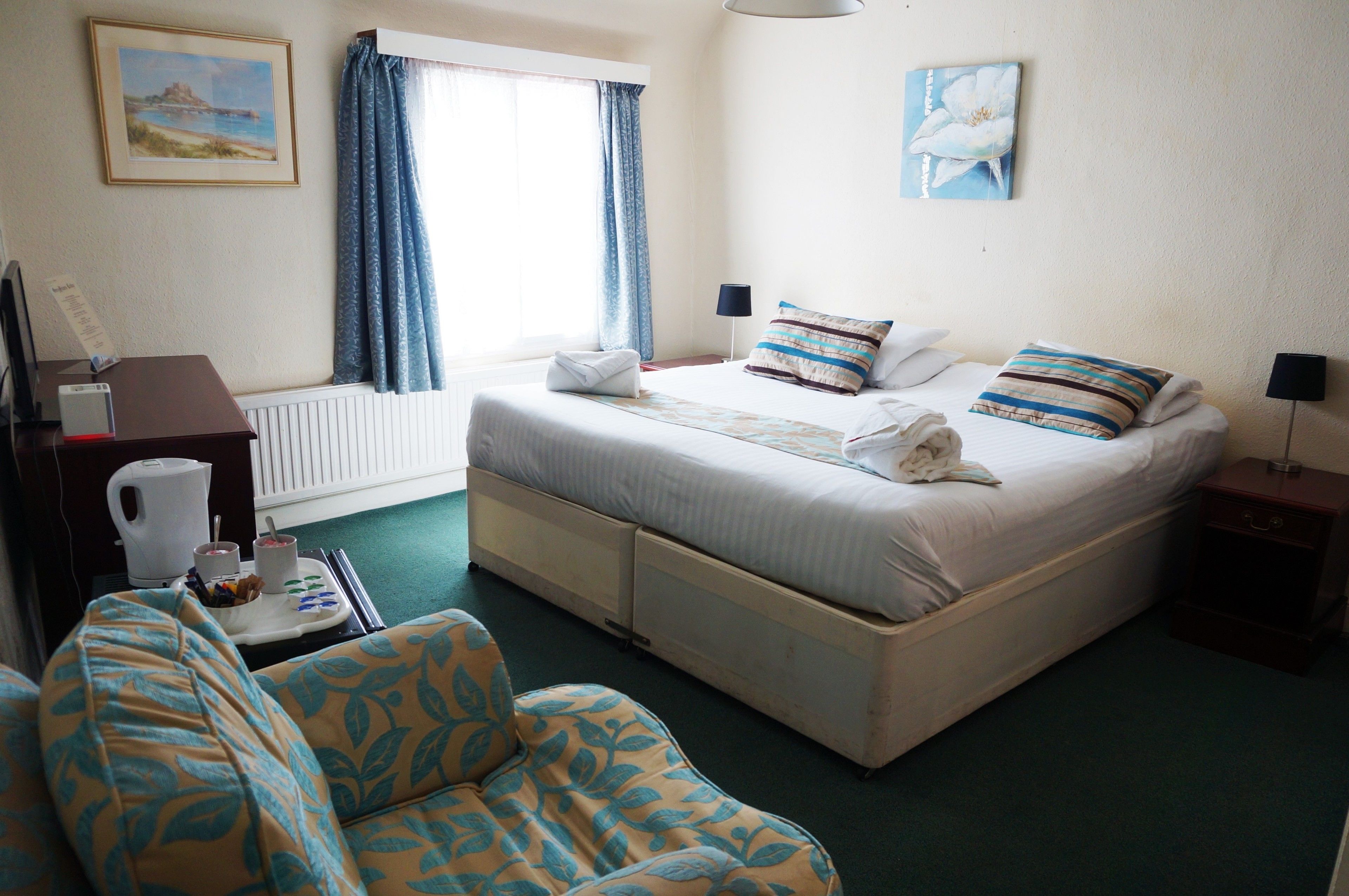 Ein Hotelzimmer mit Bett, Sessel, kleinem Tisch mit Wasserkocher und Tassen sowie einem Fenster mit Vorhängen.