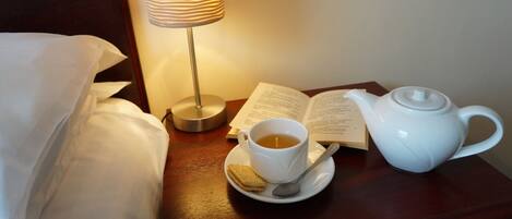 Een keurig opgemaakt bed met witte lakens, een nachtkastje met een witte theepot, een kop thee, een koekje, een open boek en een lamp.