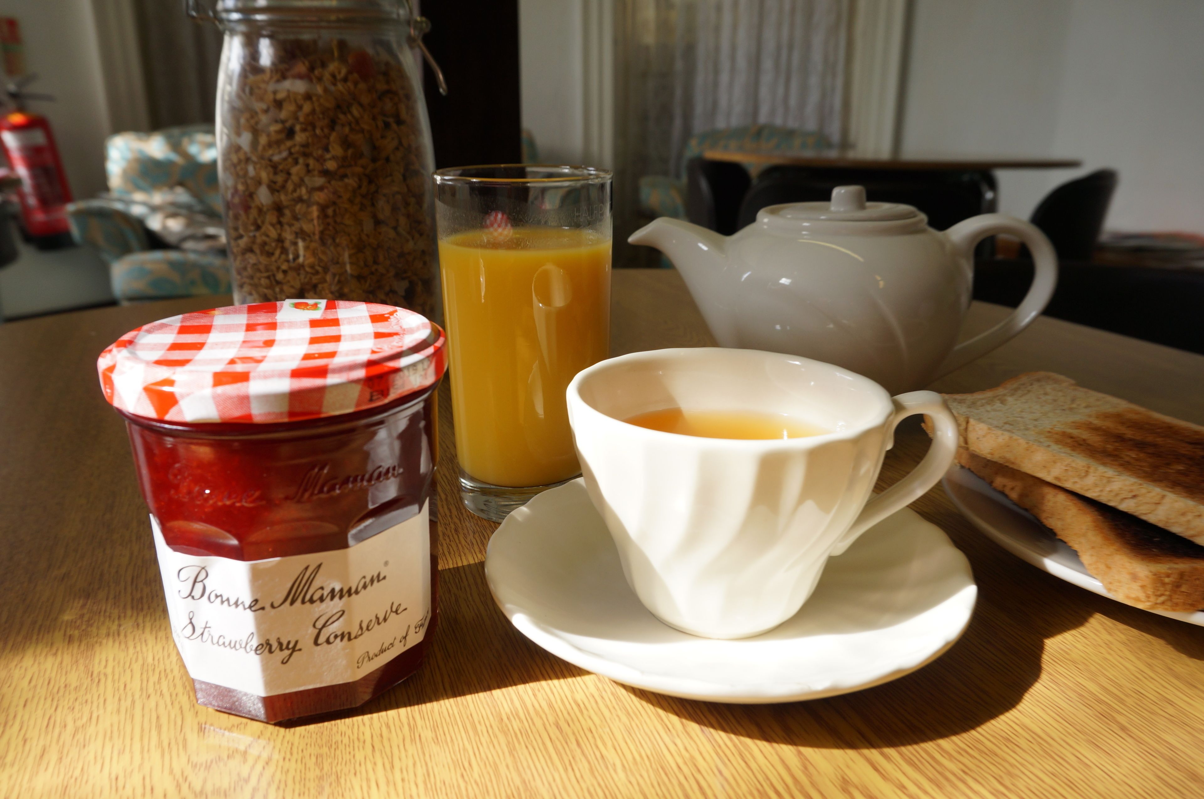 A breakfast setting with a jar of Bonne Maman strawberry conserve, a glass of orange juice, a teapot, and toast on a wooden table.
