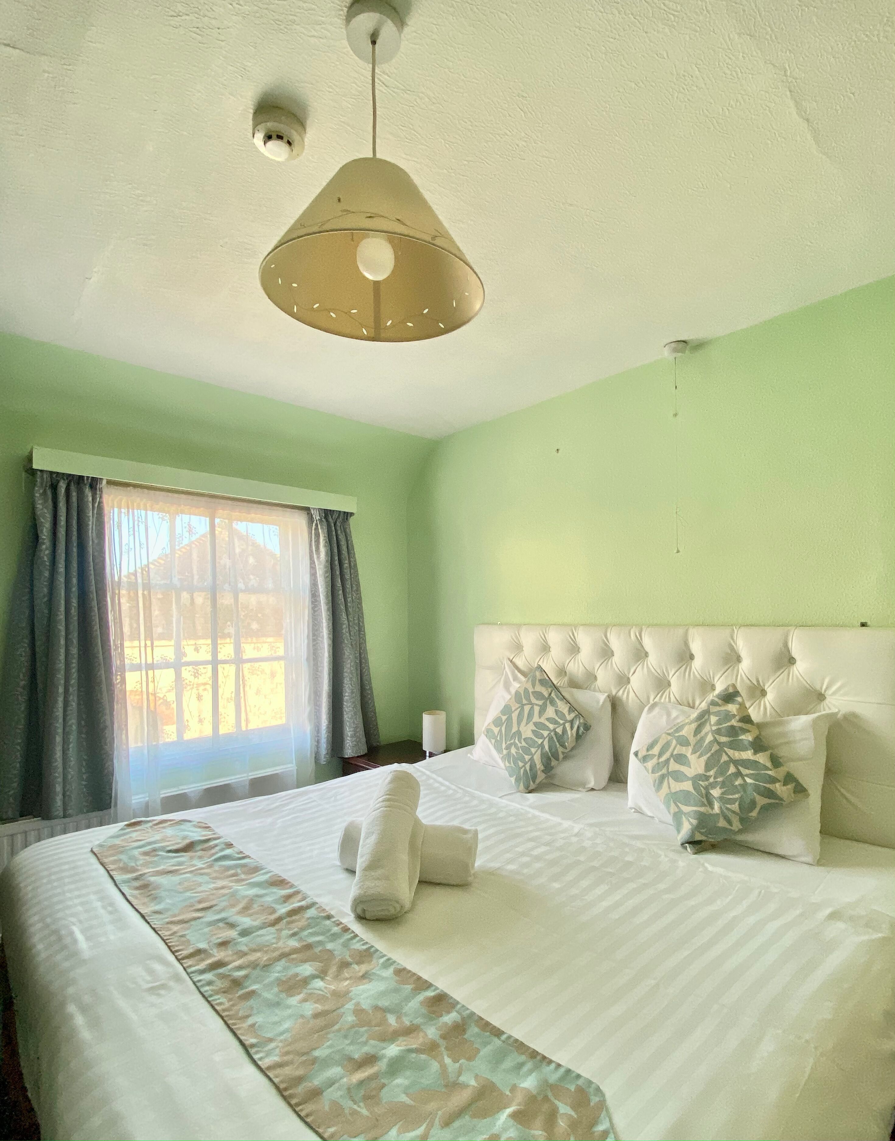 Una cama bien tendida con edredón blanco, dos almohadas y una toalla enrollada. Se ve una ventana con cortinas, y un aplique en el techo sobre la cama.