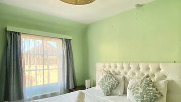 Una cama bien tendida con edredón blanco, dos almohadas y una toalla enrollada. Se ve una ventana con cortinas, y un aplique en el techo sobre la cama.
