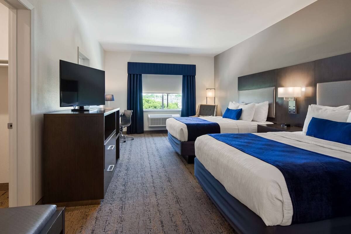 A hotel room with two beds, a desk with a TV, a lamp, and a view of greenery outside.