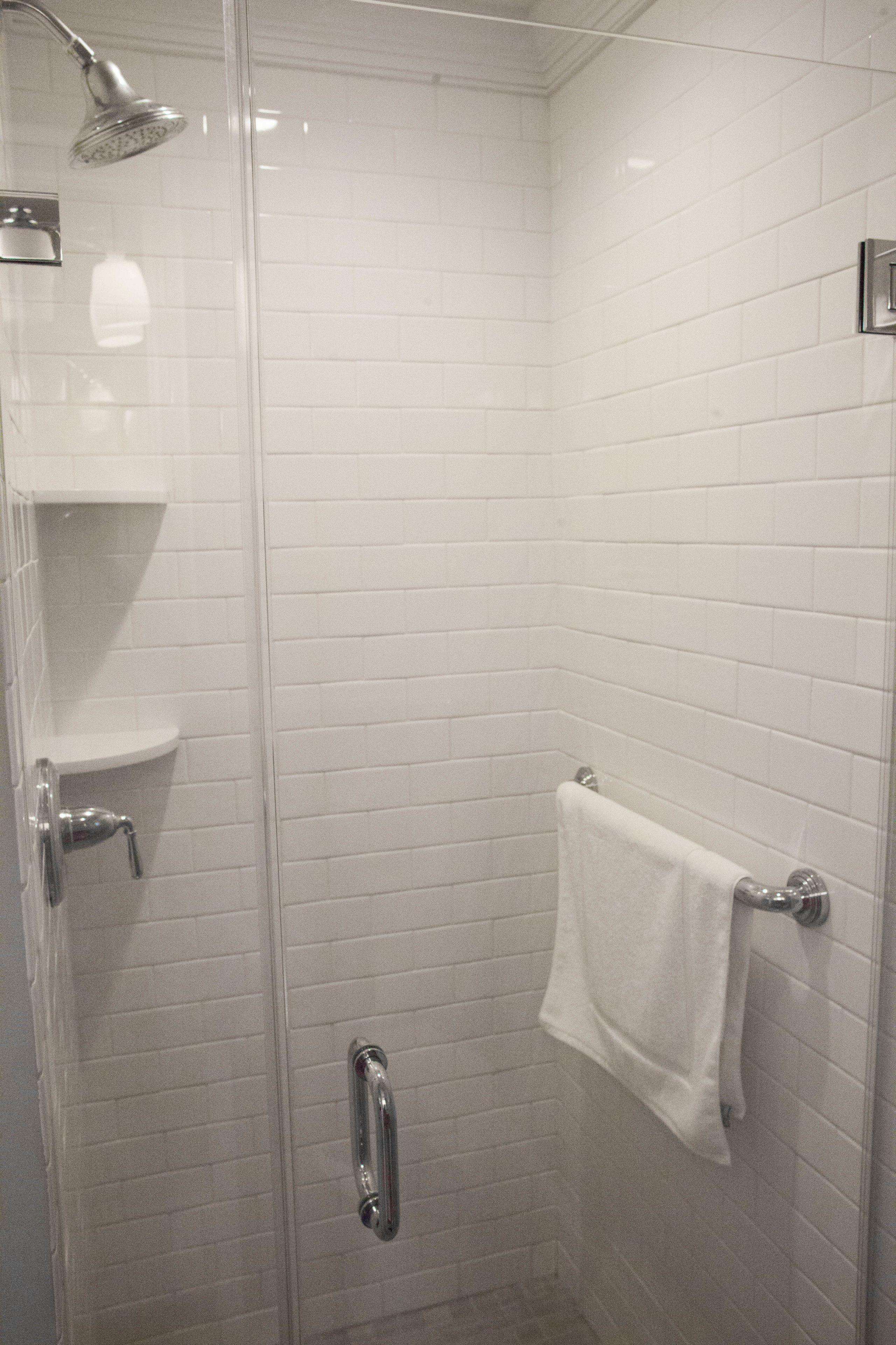 Una ducha con paredes de cristal, azulejos blancos, un perchero y un cabezal de ducha cromado.