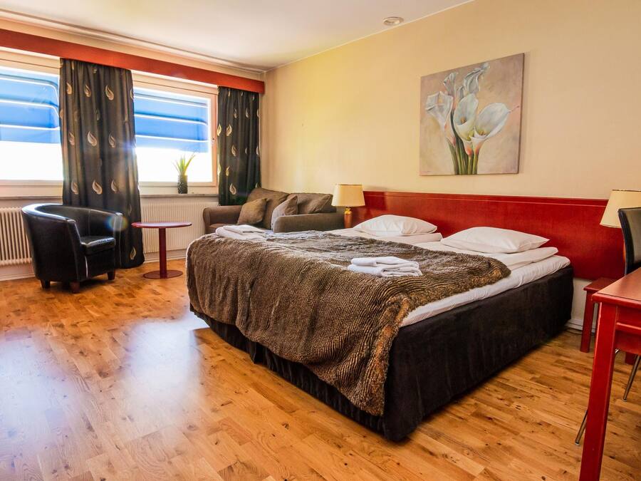 Ett hotellrum med en stor säng, ett litet bord, en stol och ett fönster med gardiner.