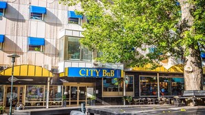 A city building with a 'CITY B&B' sign, outdoor seating, and a large tree in front. - Skoogs BnB (Pitea)
