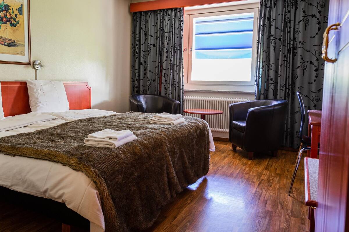 Een hotelkamer met een bed, een stoel, een klein tafeltje en een raam met gordijnen.
