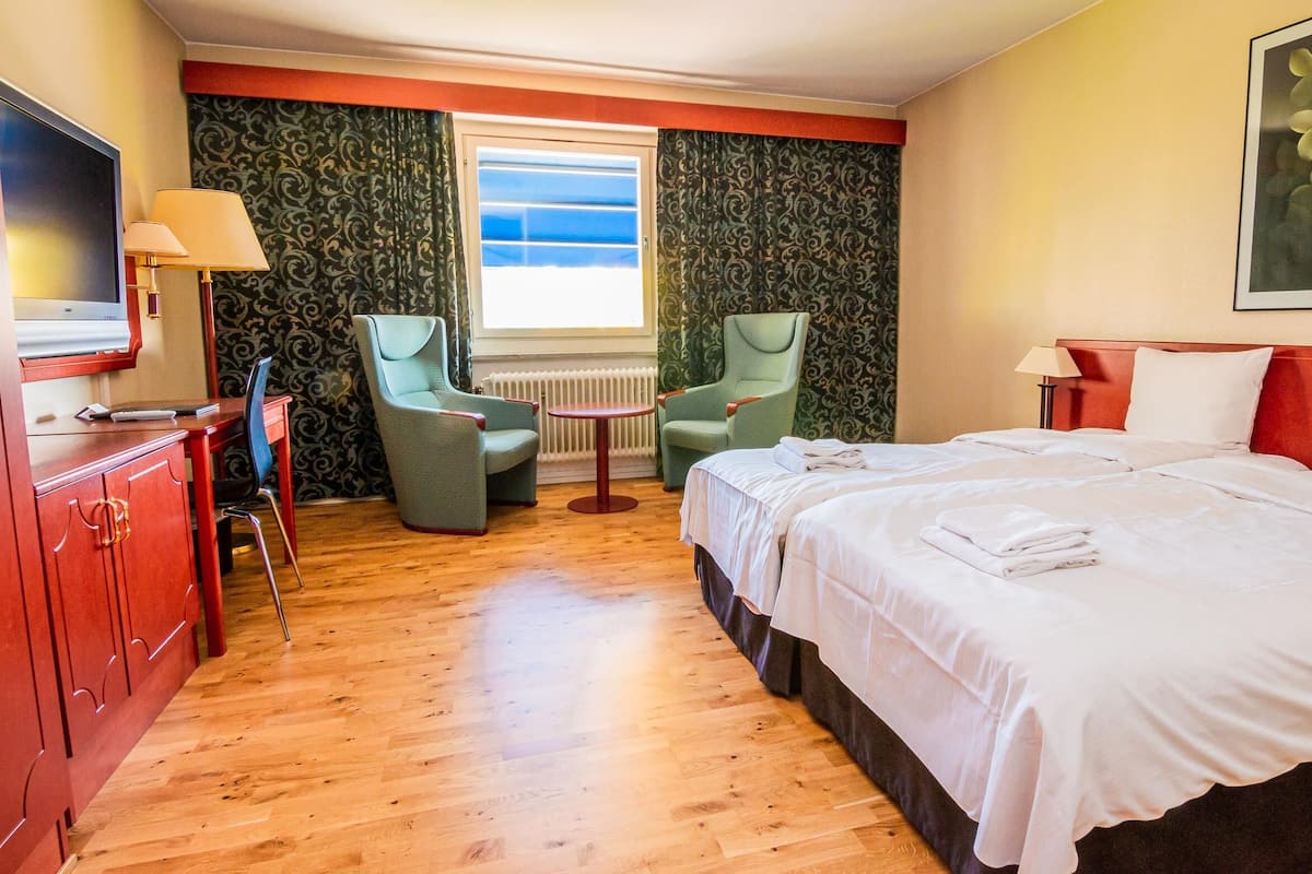 Een hotelkamer met een bed, twee fauteuils, een bureau met lamp, een televisie en een raam met gordijnen.