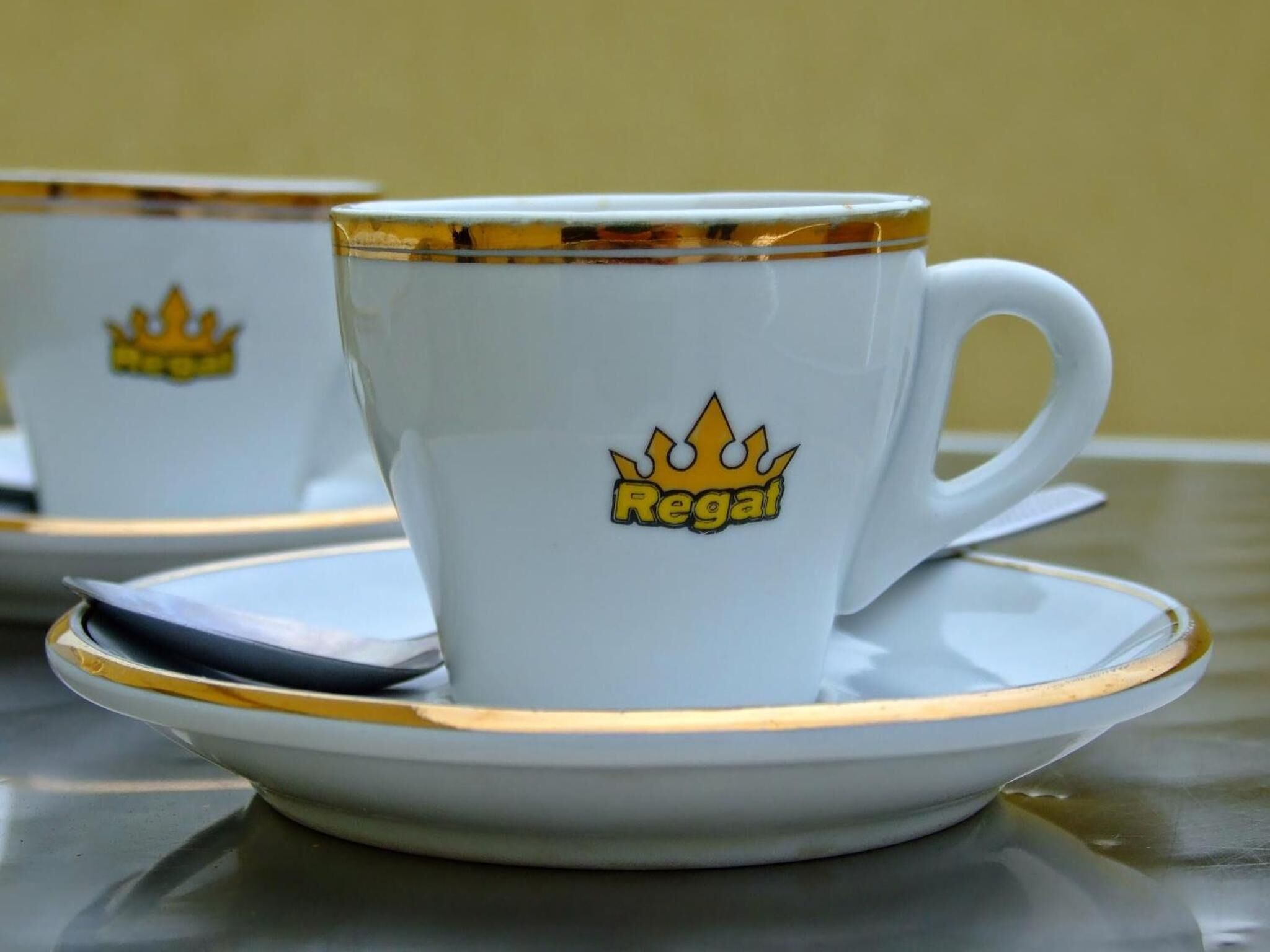 A white cup with a gold rim and a gold crown logo, placed on a matching saucer.