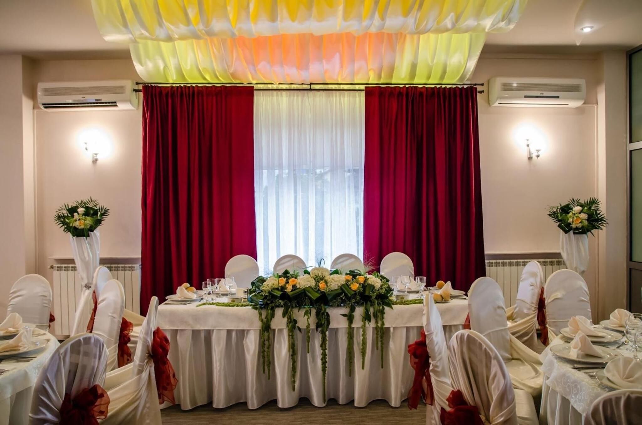 Une salle de banquet où les tables sont dressées pour un événement formel, avec des nappes blanches, des rideaux rouges et des décorations florales au centre.