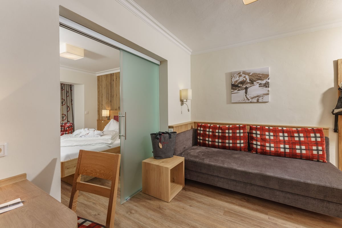 A hotel room with a wooden floor, a sofa with red and white checkered pillows, a wooden nightstand, a small table, a chair, a sliding door, a wall-mounted lamp, and a framed picture on the wall.