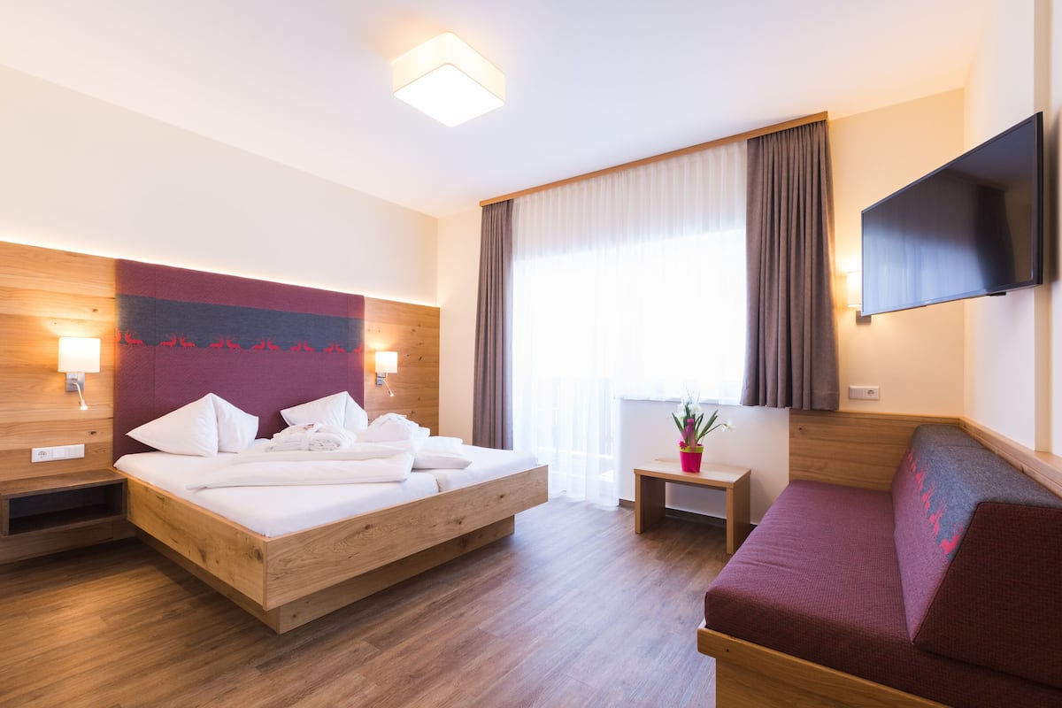 A hotel room with a wooden bed, a sofa, a TV, a small table with a plant, and a window with curtains.