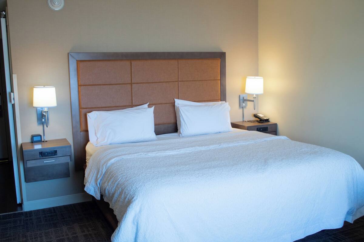 Una habitación de hotel con una cama grande, dos mesitas de noche con lámparas, un tablero auxiliar y un reloj de pared.