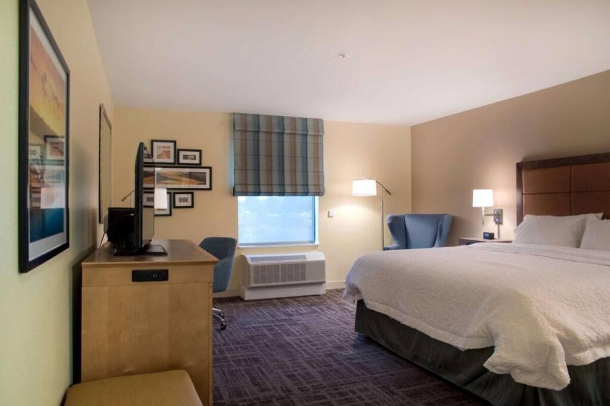 Habitación de hotel con cama, escritorio, silla, ventana con persianas y televisor instalado en la pared.