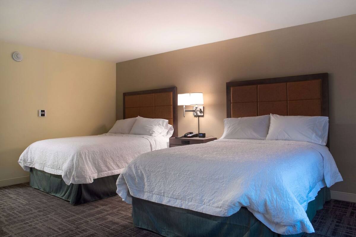 A hotel room with two beds, a bedside table with a lamp, and a wall-mounted phone.