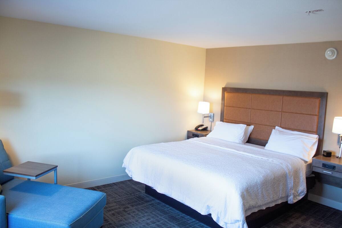 Habitación de hotel con una cama grande, dos mesitas de noche, una silla y un tocador con lámpara.