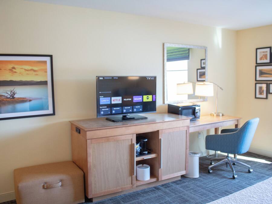 Une chambre d’hôtel avec un bureau en bois, un téléviseur affichant l’interface d’un service de diffusion en continu, une lampe, une chaise, une valise et un tableau encadré au mur.