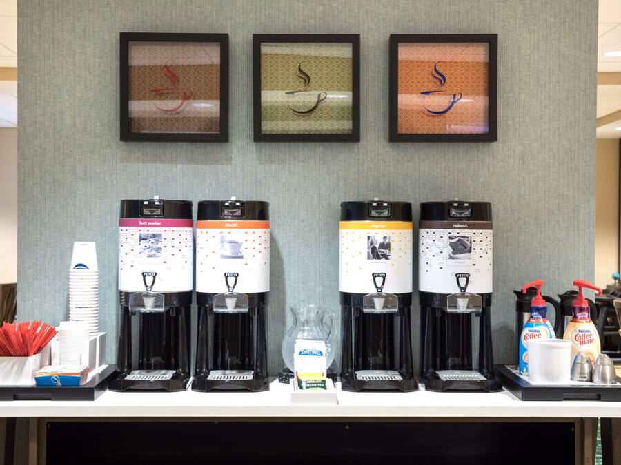 A coffee station with four dispensers, a hot water dispenser, and a coffee maker. There are framed artworks on the wall above.