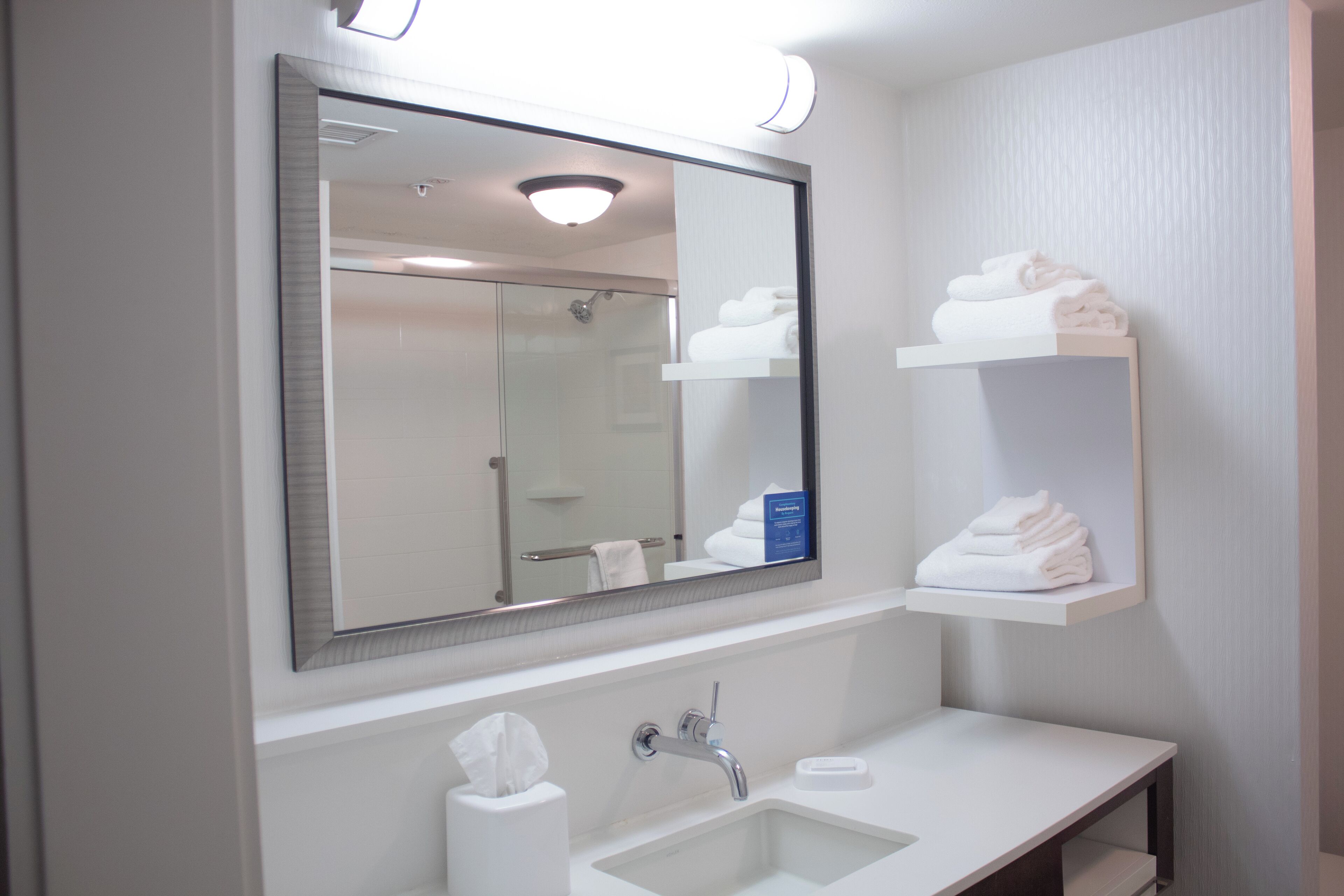 A bathroom with a large mirror, a sink, and shelves with folded towels.