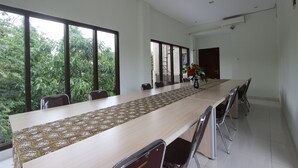 A long conference table with a patterned table runner, surrounded by chairs, in a room with large windows and a view of greenery outside. - RedDoorz @ Turangga Sari (Depok)