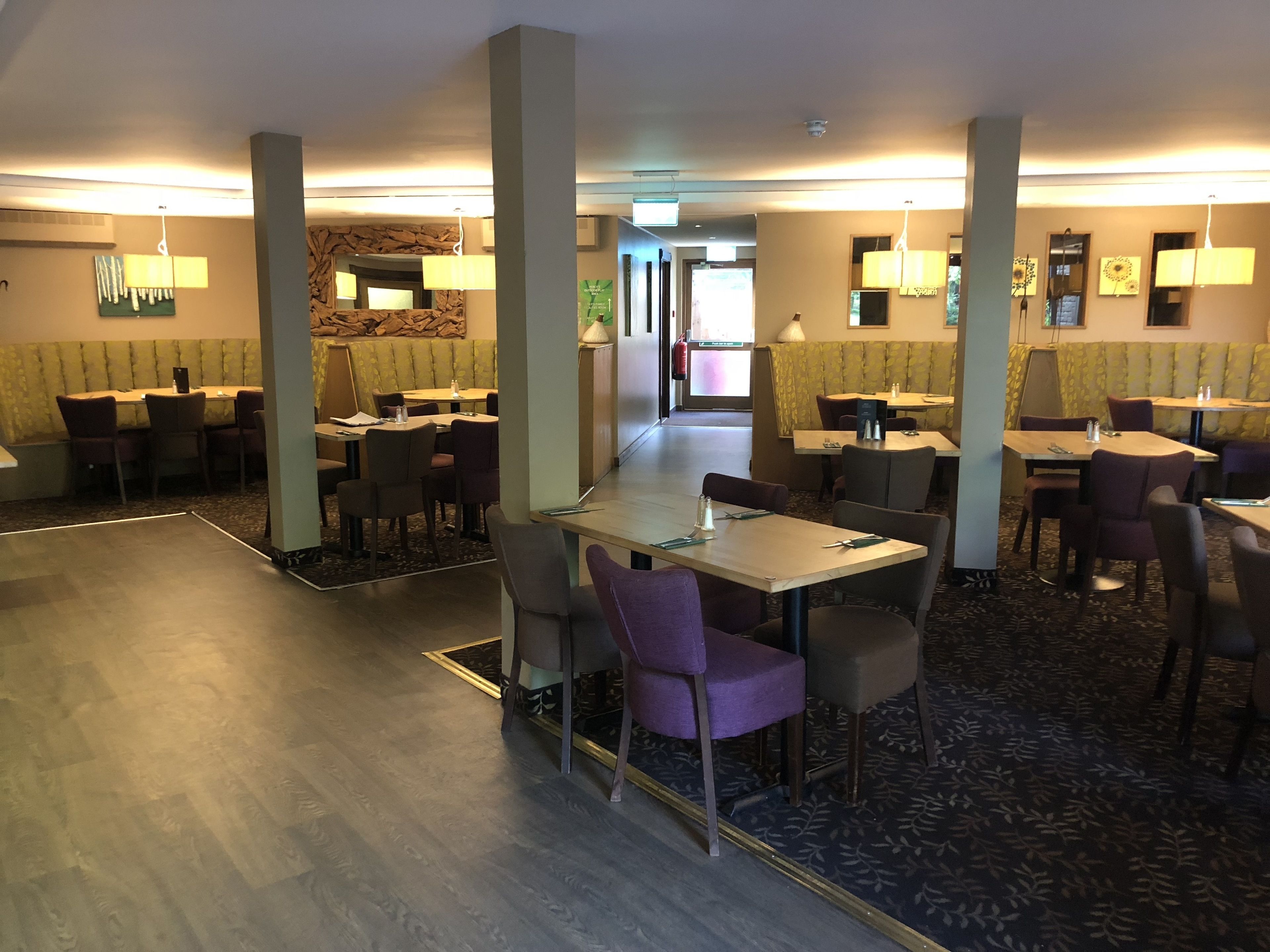 A hotel dining area with multiple tables and chairs, a patterned carpet, and wall-mounted lamps.