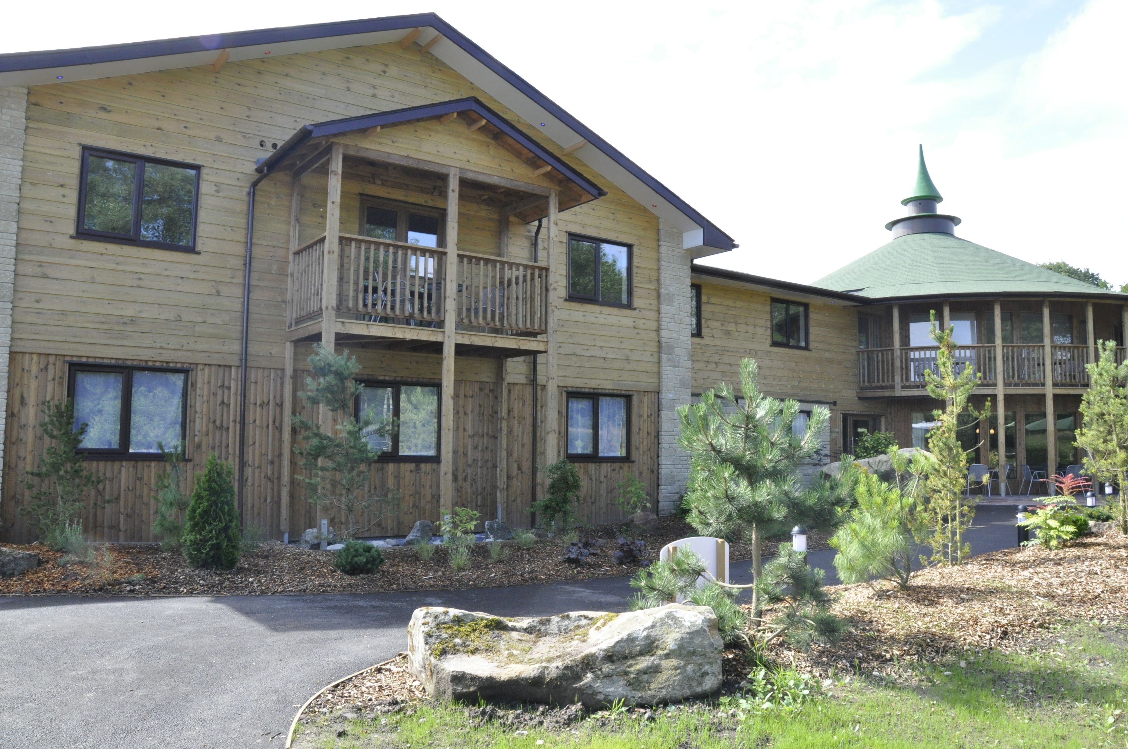 A two-story wooden building with a balcony on the second floor, a green-roofed structure to the right, and a landscaped front yard with rocks and small trees.