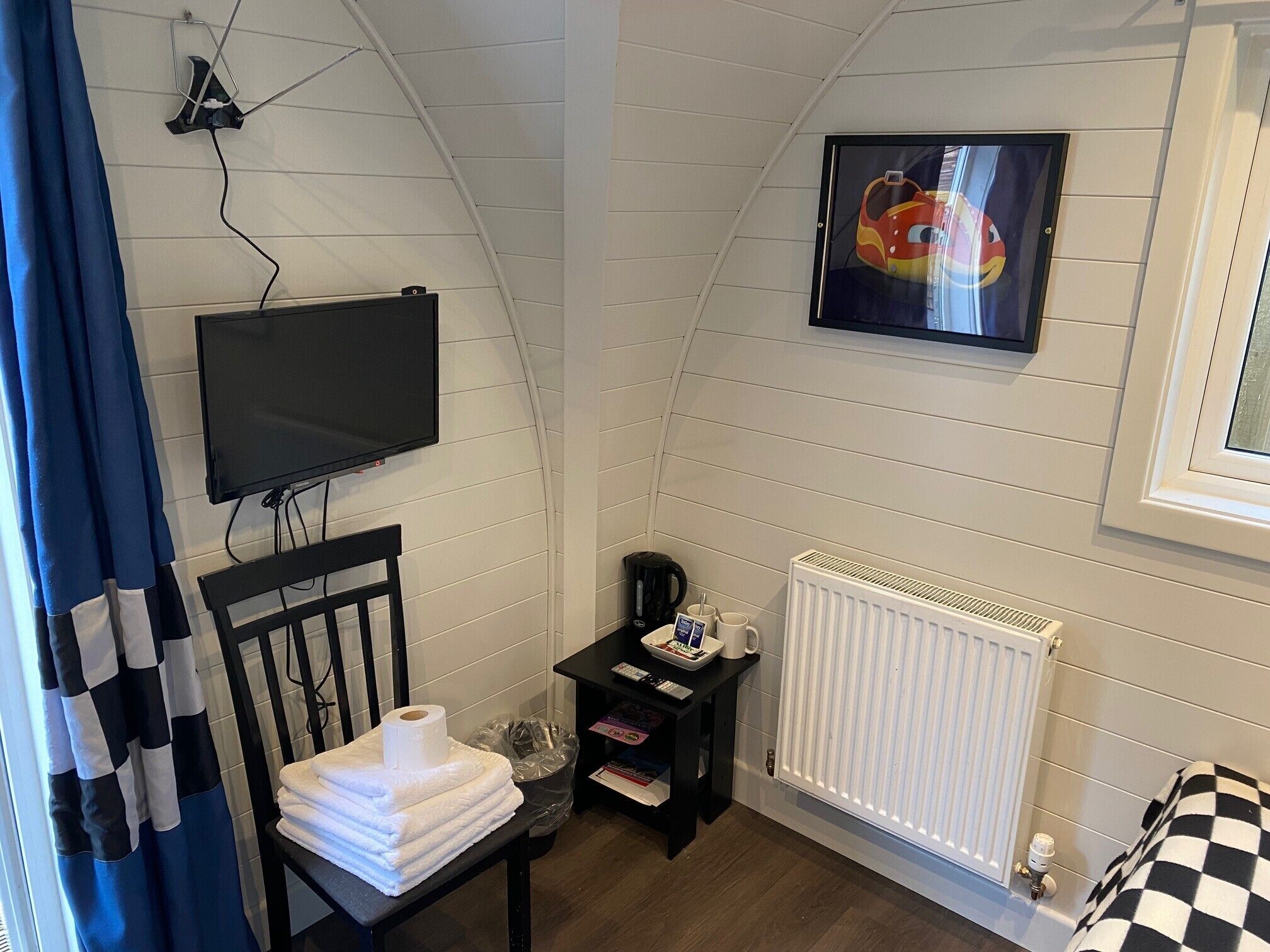 A small, cozy room with a television, a radiator, a chair, and a window with a checkered curtain.