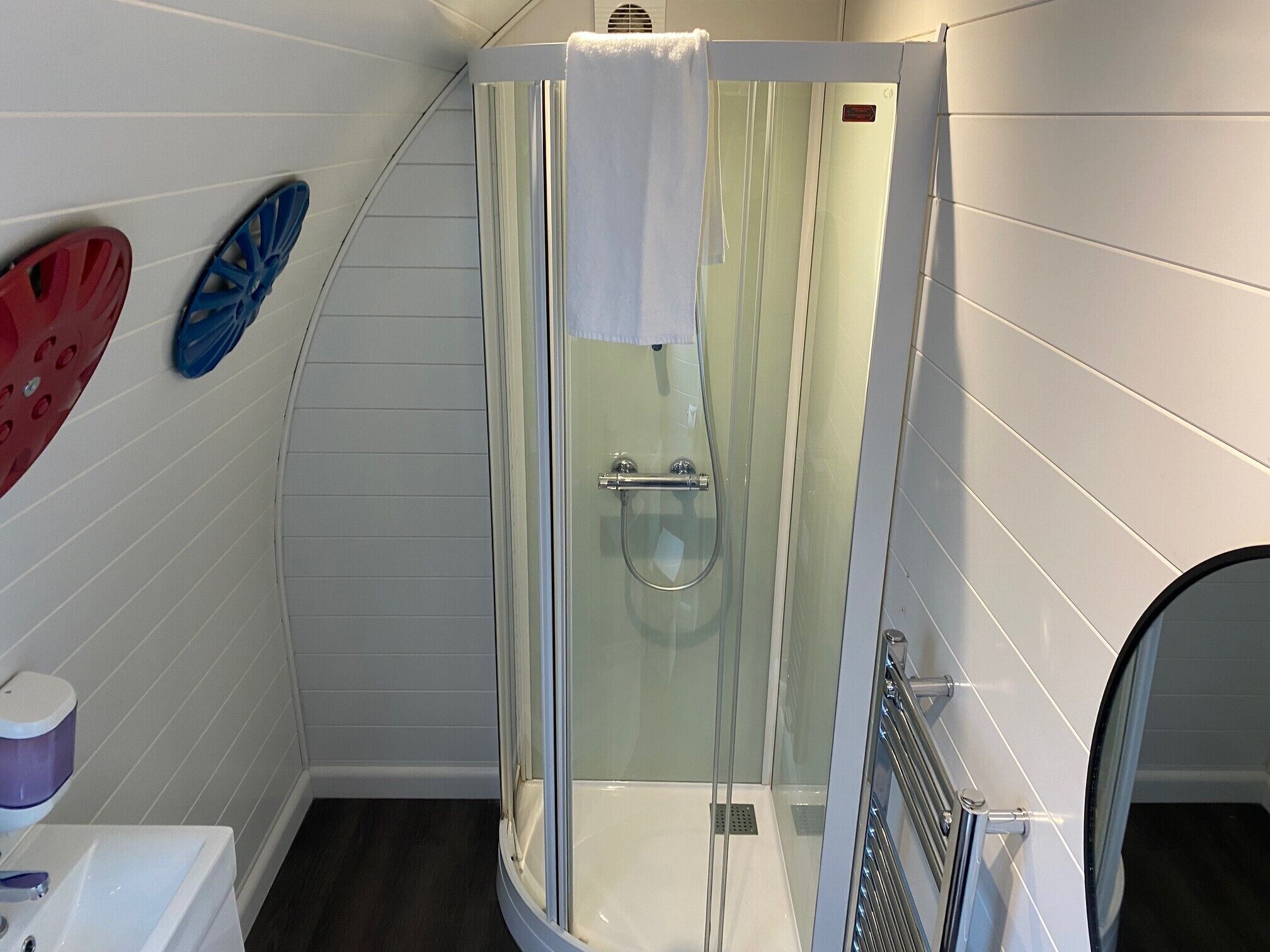 A shower with a glass door, a white towel hanging on the door, and a white sink with a purple soap dispenser.