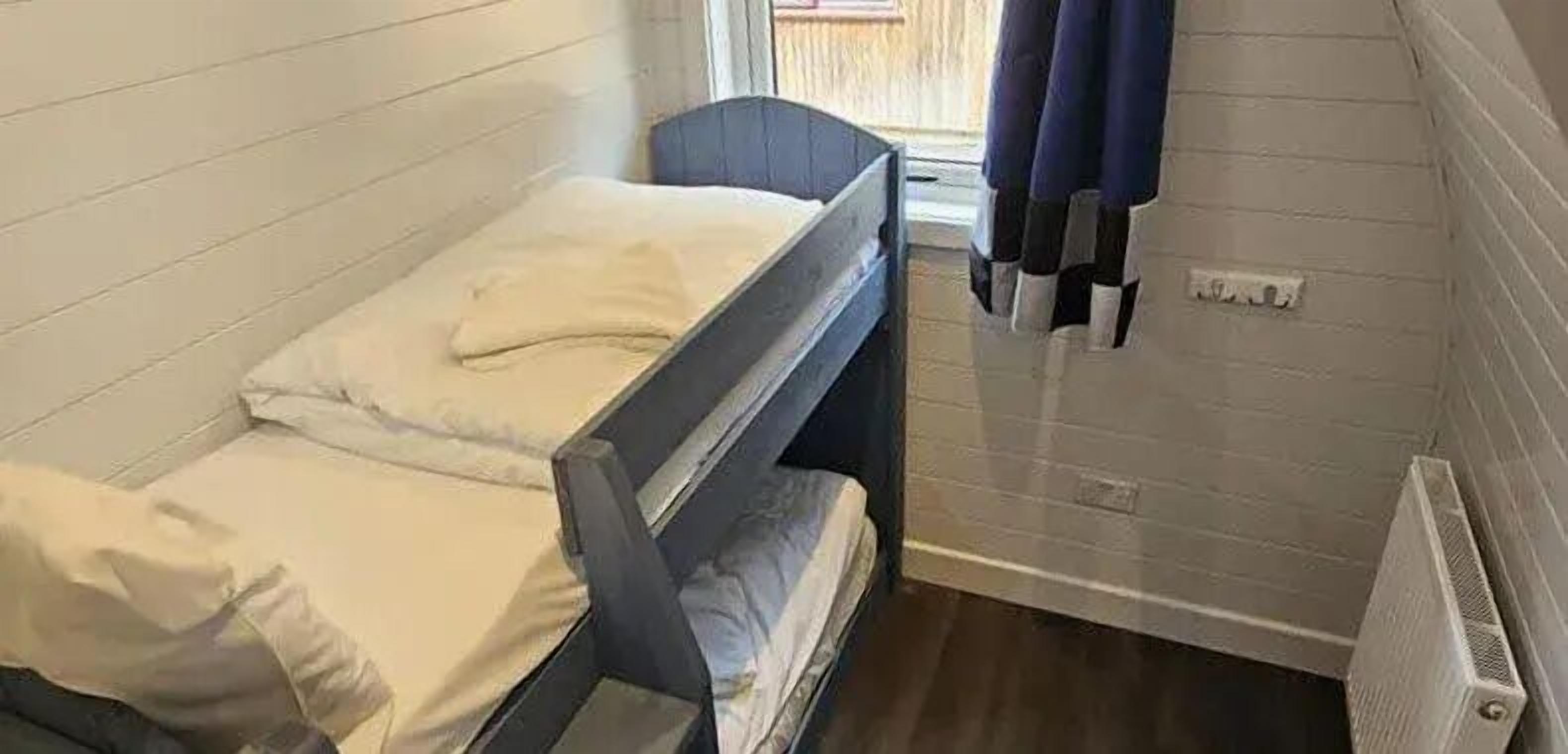 Una habitación pequeña con cama individual y literas, una ventana con cortinas y un radiador en la pared.