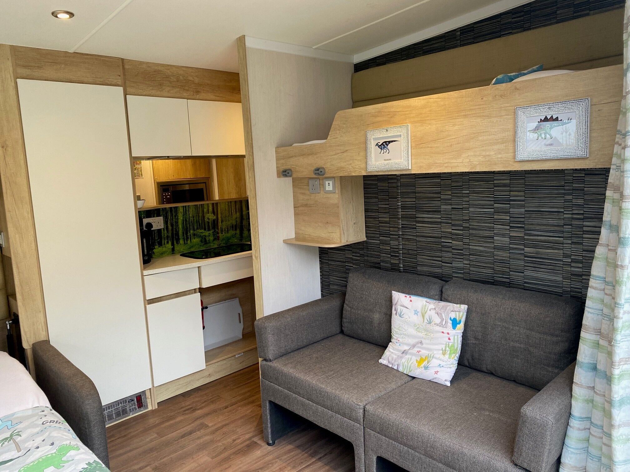 A compact living space with a kitchenette, a sofa with a dinosaur-themed pillow, and a wall-mounted shelf with framed pictures.