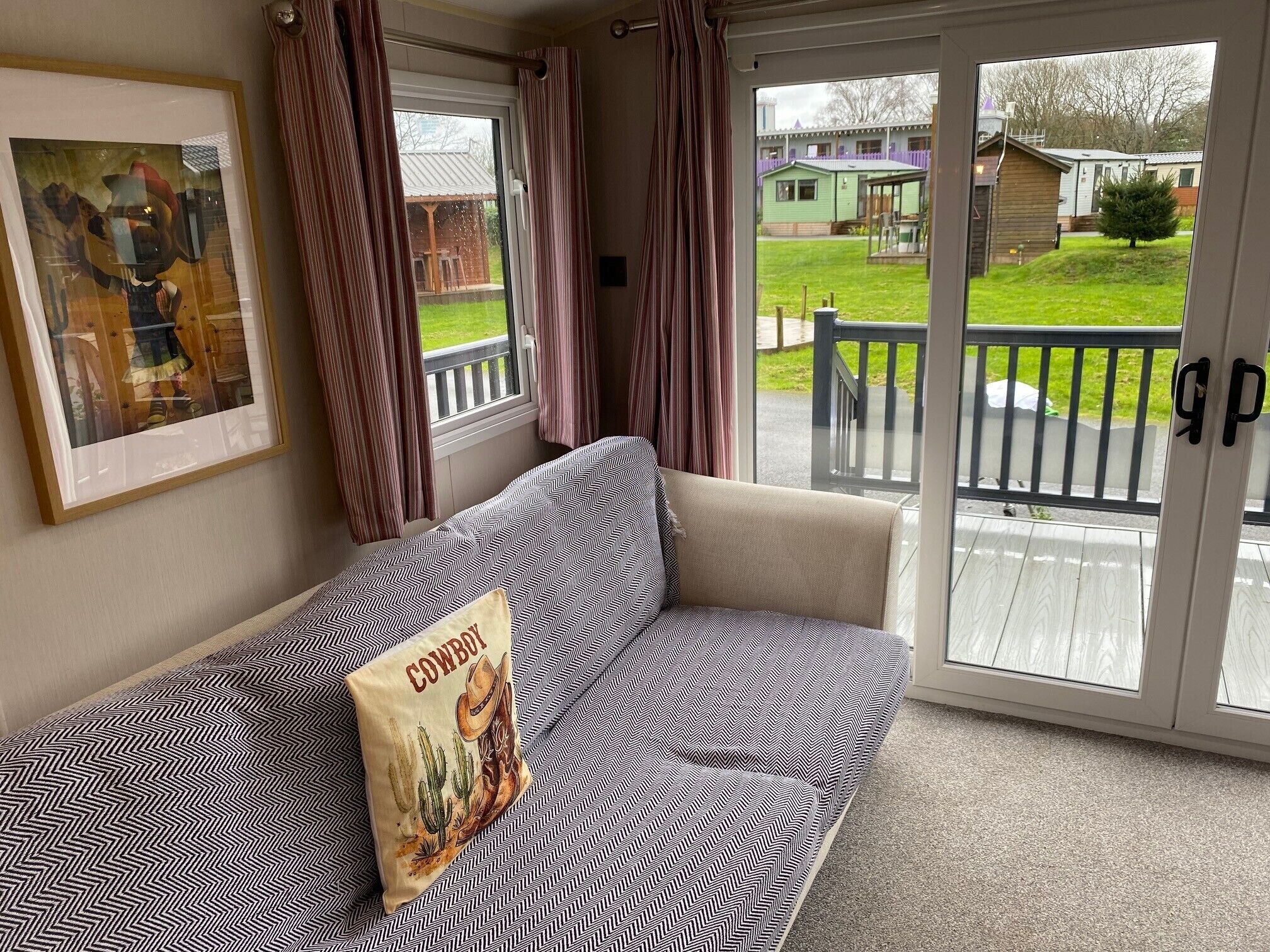 A room with a sofa, a framed picture, and a sliding glass door leading to an outdoor area with a deck and greenery.