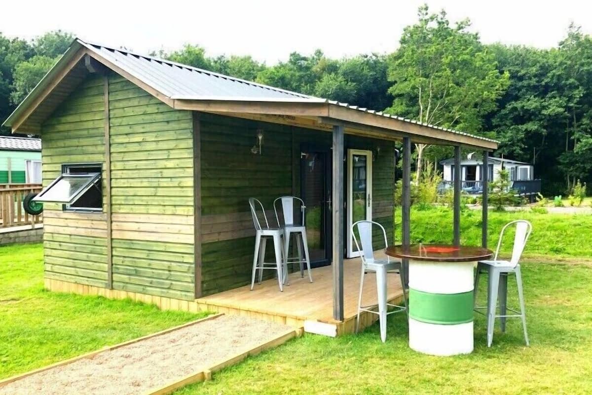 Una pequeña cabaña de madera con un porche, dos sillas, una mesa y una parrilla.