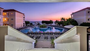 A hotel pool area with multiple lounge chairs and a central water feature, surrounded by buildings and palm trees. - Porto Iliessa by GHH (Zakynthos)
