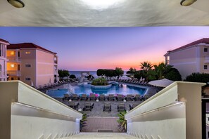 A hotel pool area with multiple lounge chairs and a central water feature, surrounded by buildings and palm trees. - Porto Iliessa by GHH (Zakynthos)
