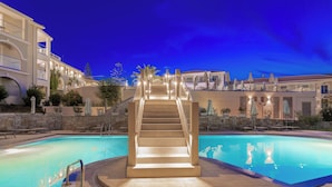 Una piscina con una escalera que lleva a un balcón, rodeada de edificios y mobiliario exterior.
