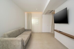 A modern living room with a beige sofa, a flat-screen TV mounted on the wall, and a wooden shelf. - Tarra Seaside Hotel (Apokoronas)