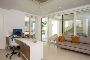 A modern office space with a desk, computer, and office chair. There is a sofa with cushions and a chair. The room has large windows and a view of the outside. - Tarra Seaside Hotel (Apokoronas)