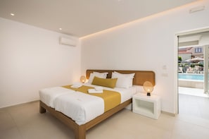 A modern hotel room with a wooden bed, white bedding, a small bedside table with a lamp, and a view of an outdoor pool area. - Tarra Seaside Hotel (Apokoronas)