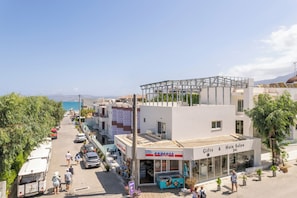 A coastal street with a 'Express Market' and 'Gifts & Hair Salon' on the ground floor, a white building with a balcony, and a clear view of the sea. - Tarra Seaside Hotel (Apokoronas)
