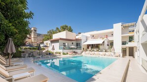 A pool area with sun loungers and umbrellas, surrounded by buildings with balconies and a hillside in the background. - Tarra Seaside Hotel (Apokoronas)