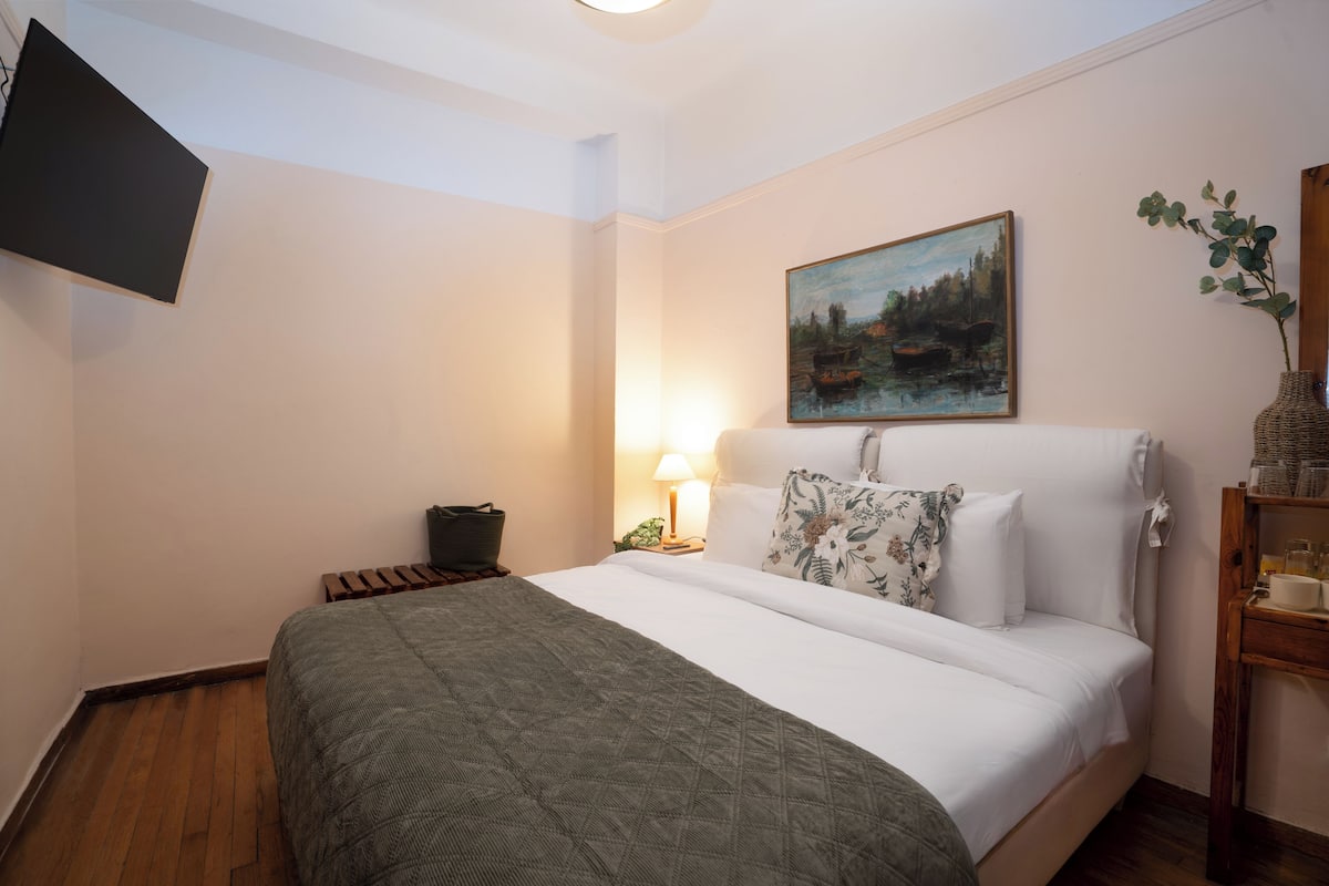 A neatly arranged hotel room with a bed, a painting, a nightstand, a lamp, a vase with plants, and a television mounted on the wall.